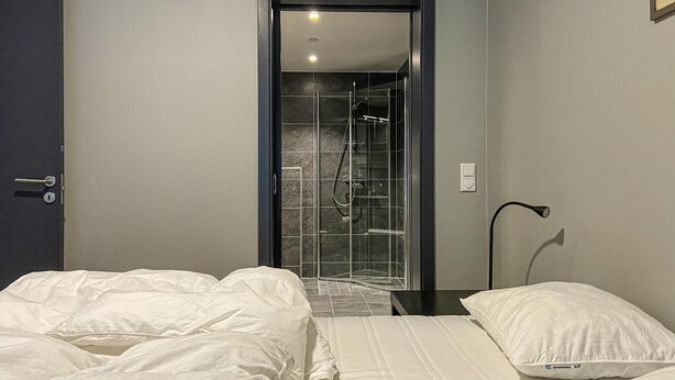 Camera d'albergo con un letto, un comodino, una lampada e un angolo doccia con porte in vetro.