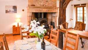 Un coin repas chaleureux avec une cheminée en pierre, des poutres en bois et une table dressée pour deux personnes, avec des assiettes et une bouteille de vin.