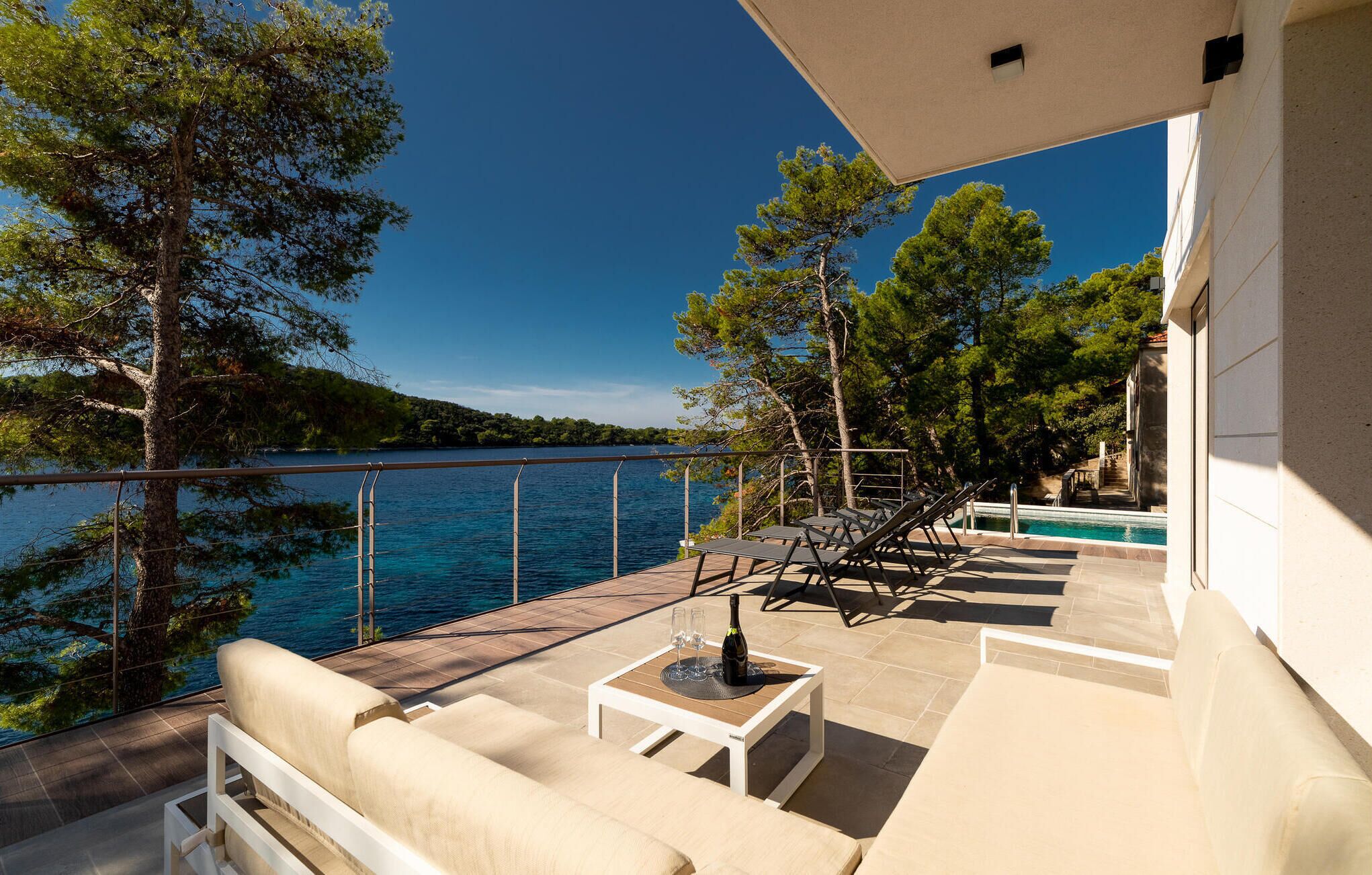 A terrace with outdoor seating, a pool, and a view of a lake surrounded by trees.