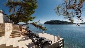 Ein Anwesen am Wasserufer mit einer Betonstiege, die zu einer Terrasse mit Liegestühlen, einem Pool und Blick auf einen See sowie eine entfernte Insel führt.