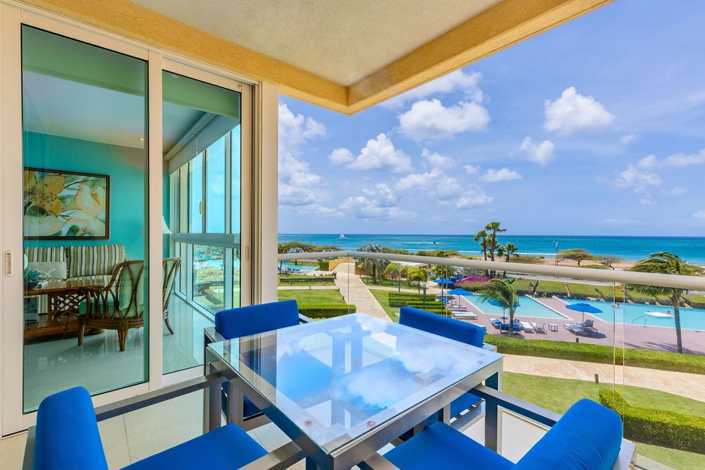 Un balcon avec une table en verre et des chaises bleues, donnant sur une piscine et une plage.