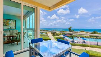 Un balcon avec une table en verre et des chaises bleues, donnant sur une piscine et une plage.