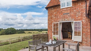 A rustic brick house with a patio table and chairs, a wooden fence, and a view of a green field.