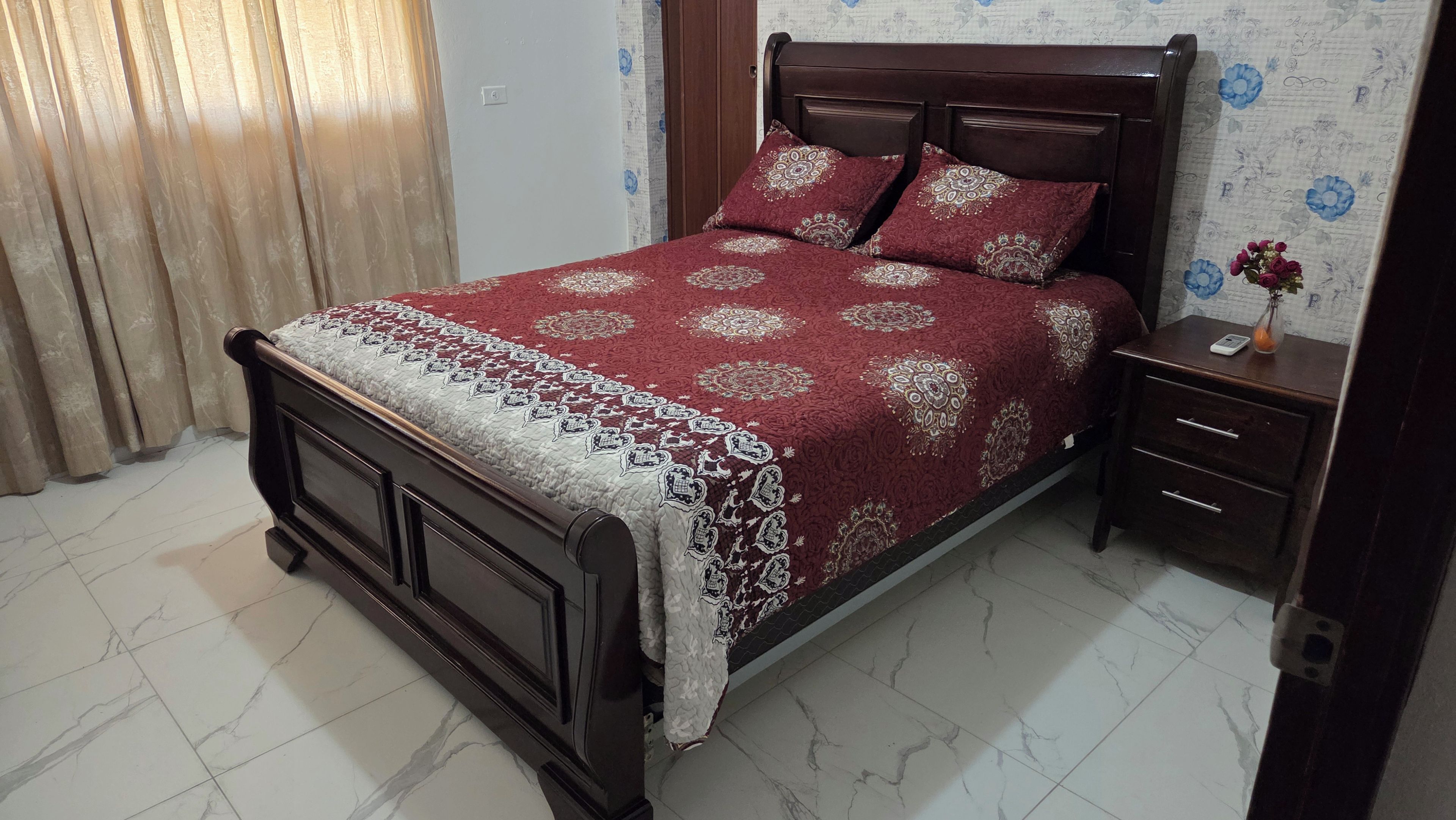 A bedroom with a wooden bed, a bedside table, a nightstand, and a wall with floral wallpaper.
