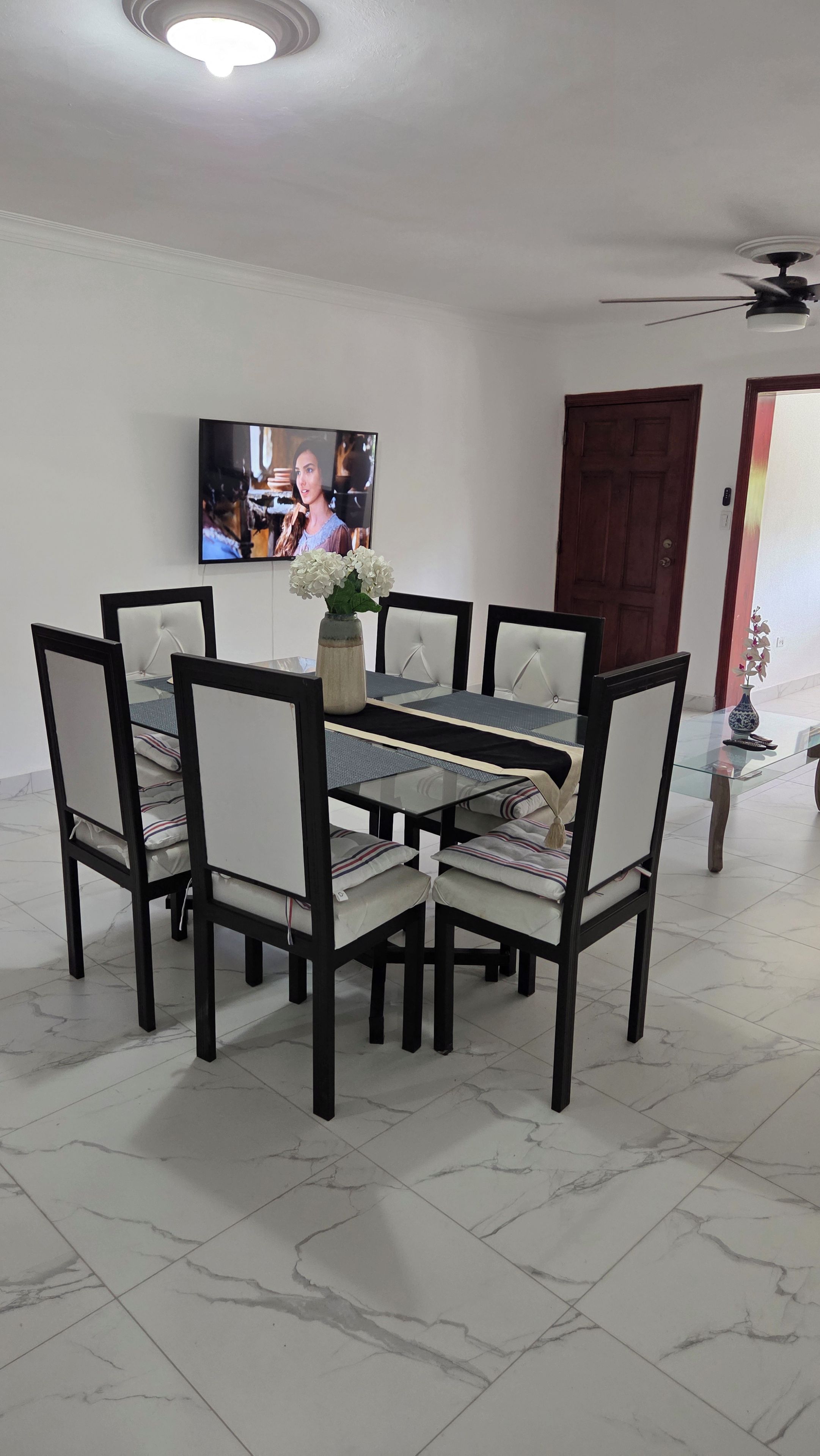 Un coin repas avec une table en verre, des chaises rembourrées, un vase avec des fleurs, un téléviseur fixé au mur et un ventilateur de plafond.