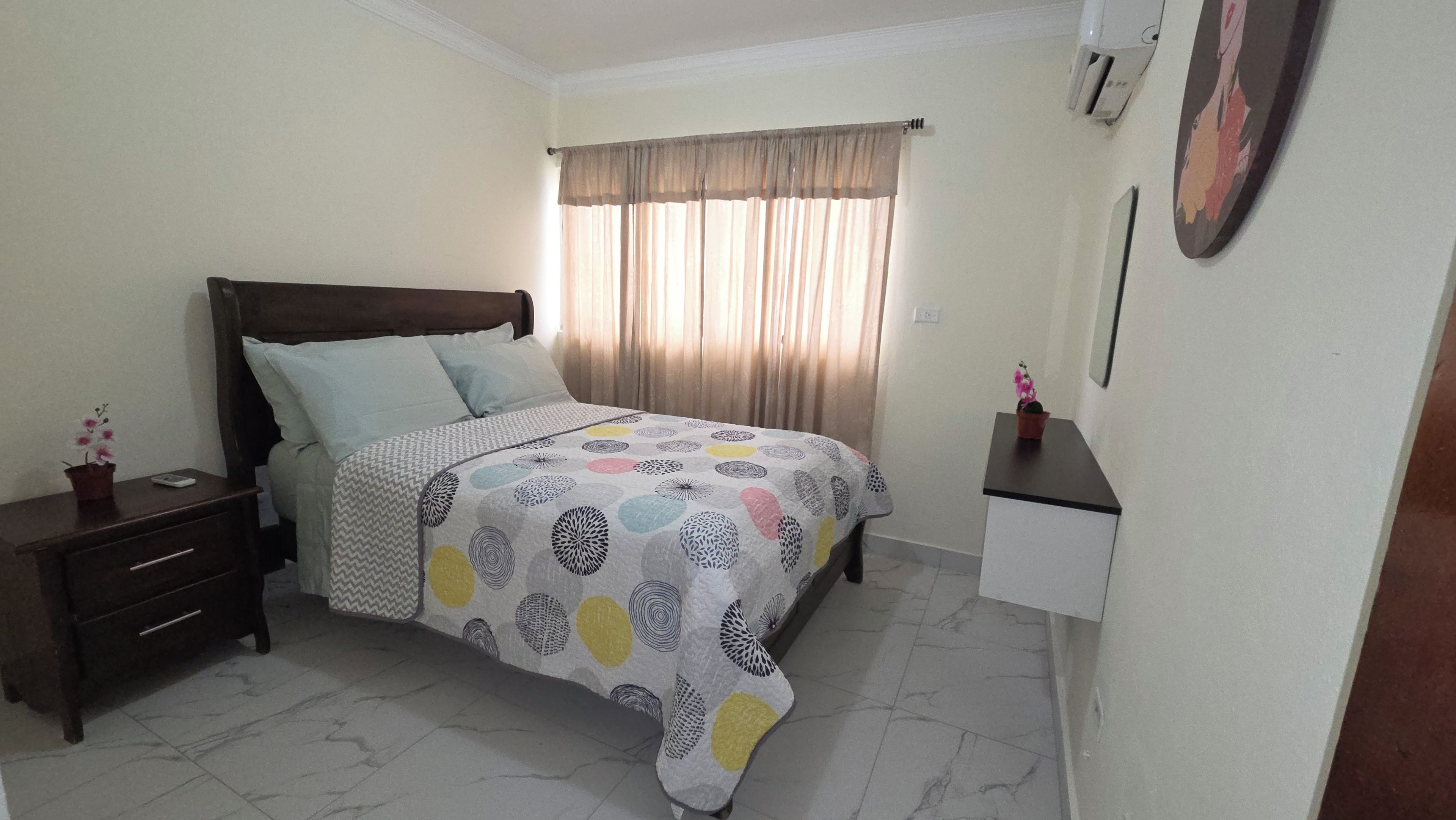 Un dormitorio con cama, mesita de noche, cómoda, ventana con cortinas y aire acondicionado de pared.