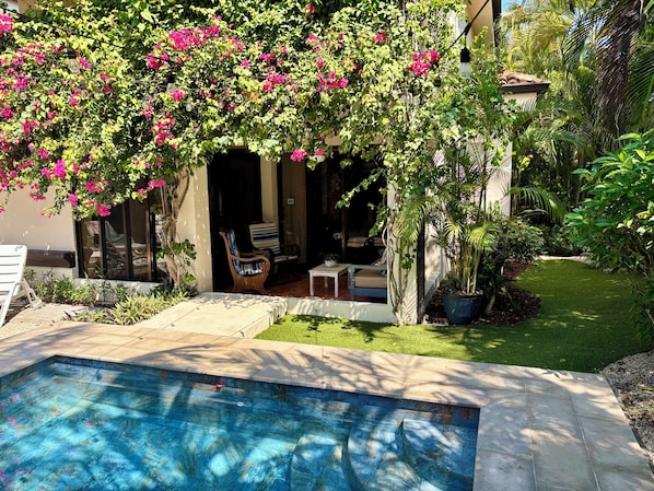 A pool with a stone border, a patio area with chairs, and a garden with flowering plants. - Tropical Jaco Beach House w/ Private Pool (Jacó)