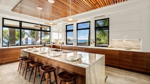 A modern kitchen with a large island, bar stools, and large windows overlooking a beach. - "Iron Man Available" Private Oceanfront Villa W/ detached Ohana and Private Pool (Kailua-Kona)