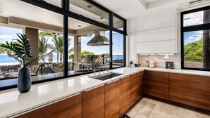 A modern kitchen with a large window overlooking a scenic view, wooden cabinets, and a sleek countertop. - "Iron Man Available" Private Oceanfront Villa W/ detached Ohana and Private Pool (Kailua-Kona)