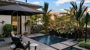 A modern outdoor pool area with a stone deck, lounge chairs, and a large umbrella. - "Iron Man Available" Private Oceanfront Villa W/ detached Ohana and Private Pool (Kailua-Kona)