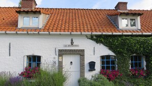 Un edificio blanco con techo de tejas rojas, una puerta blanca y dos ventanas con marcos azules. Un letrero sobre la puerta dice "LA QUITAPENAS".