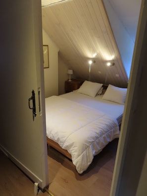 A bedroom with a sloped ceiling, a bed with white bedding, a nightstand with a lamp, and a wooden floor.