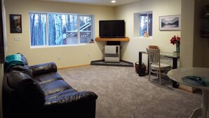 A living room with a leather couch, a television, a dining table with chairs, and a window with a view of a wooden deck and trees. - Two bedroom, fully furnished apartment with private entrance on 3 wooded acres (Fairbanks)