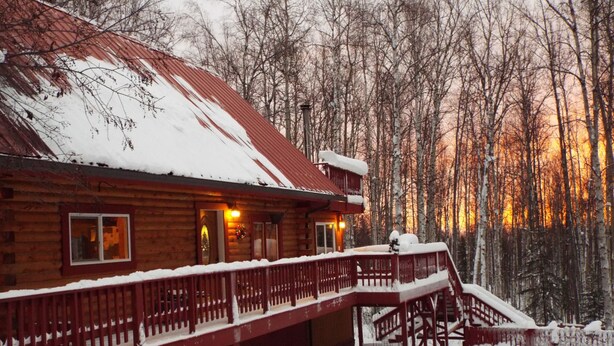 A log cabin with a wooden deck and a staircase leading to the entrance. - Two bedroom, fully furnished apartment with private entrance on 3 wooded acres (Fairbanks)