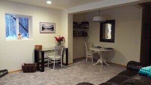 A living room with a brown leather couch, a white dining table with chairs, a small white table with a vase of red flowers, a bookshelf, and a window with a view of trees. - Two bedroom, fully furnished apartment with private entrance on 3 wooded acres (Fairbanks)