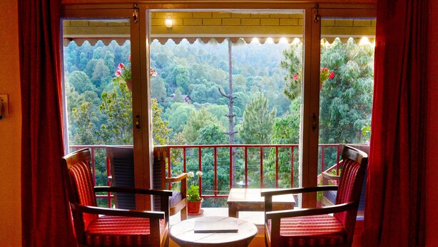 A room with two chairs and a table, a window with a view of a forest, and a door with a view of a balcony. - Majkhali Woods, Ranikhet, By Himalayan Eco Lodges (Ranikhet)