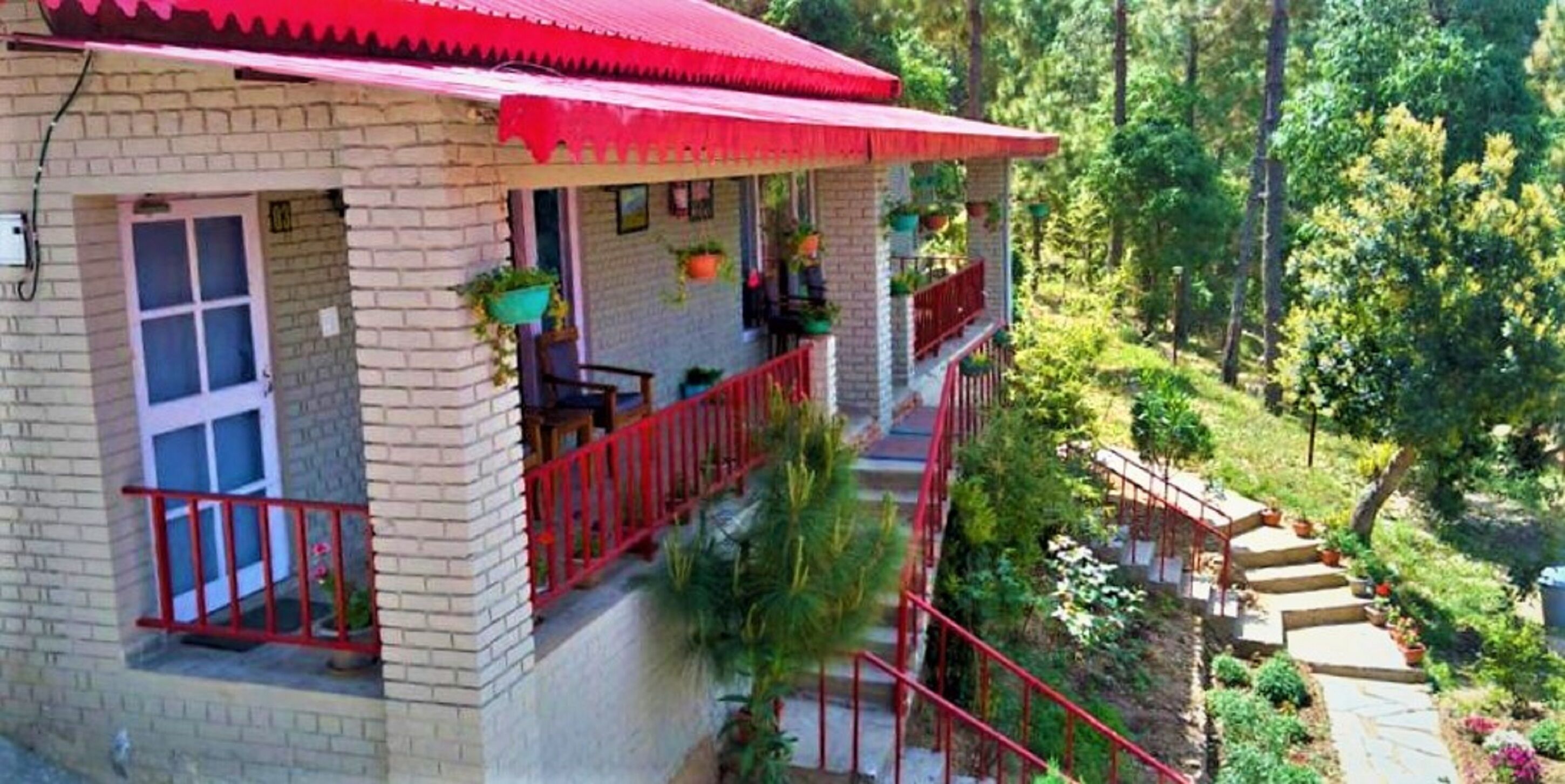 A two-story building with a red roof and balcony, surrounded by lush greenery.