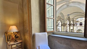 A room with a white chair by a window, a wooden cabinet with a lamp, and a view of an architectural building.