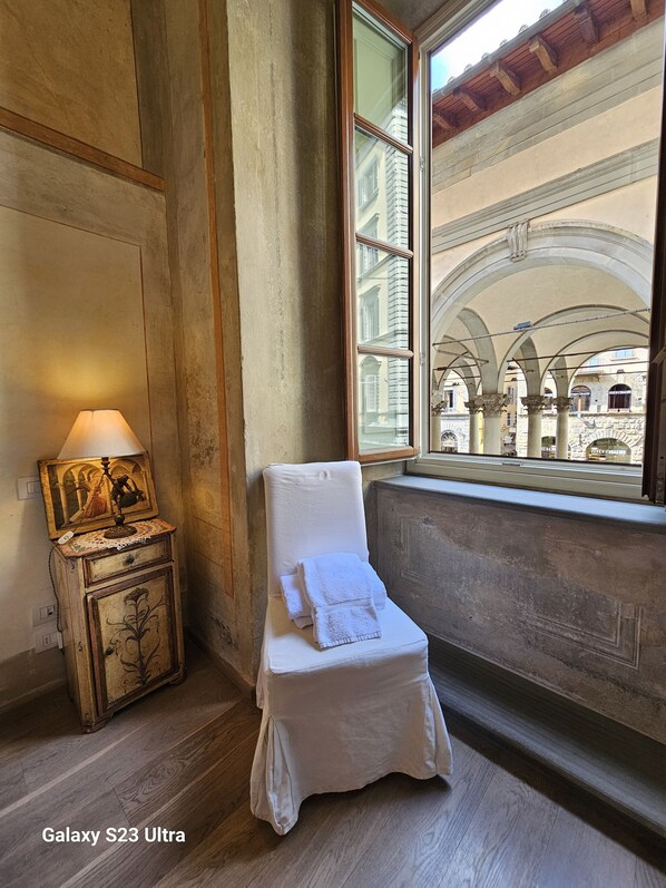 A room with a white chair by a window, a wooden cabinet with a lamp, and a view of an architectural building. - PonteVecchio Signoria Apartment (Firenze)