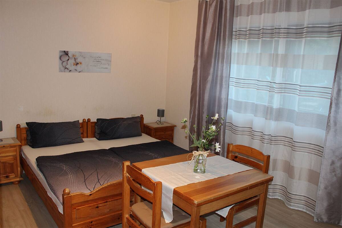 A bedroom with a double bed, a bedside table with a lamp, a wooden chair, a small table with a vase, and a window with curtains.