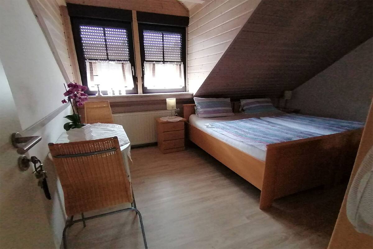 A small, cozy room with a sloped ceiling, a single bed, a chair, a small table, and a window with blinds.