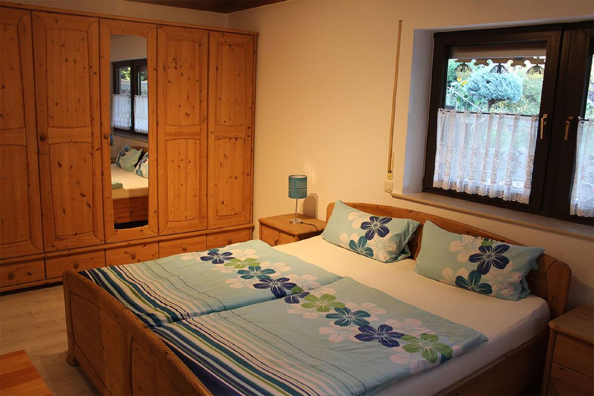 A bedroom with a wooden wardrobe, a bed with floral bedding, a nightstand with a lamp, and a window with lace curtains.