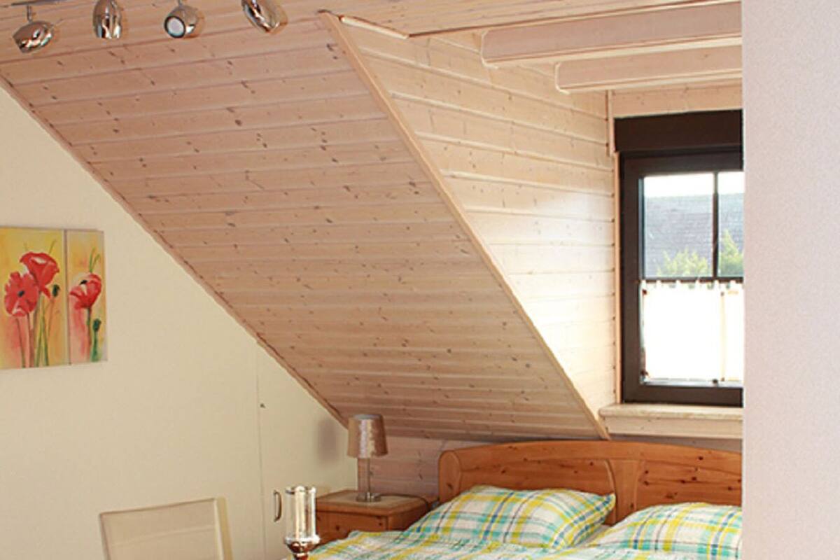 A bedroom with a wooden ceiling, a bed with plaid bedding, a table with chairs, and a window.