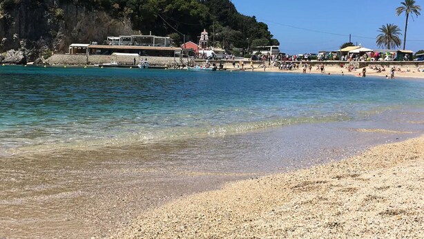 Ein Strand mit klarem Wasser, Kies und eine felsige Hügelkuppe mit Gebäuden.