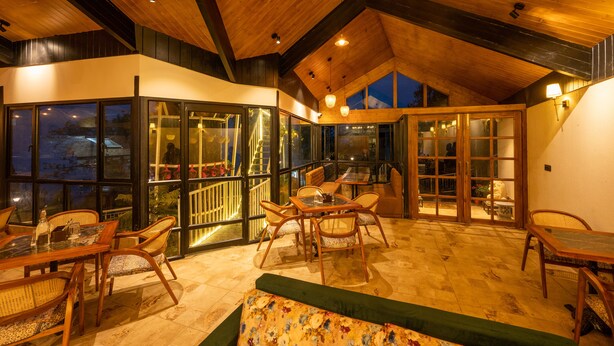 A spacious indoor area with wooden ceilings and large windows. There are multiple seating arrangements including a table and chairs, and a sofa.