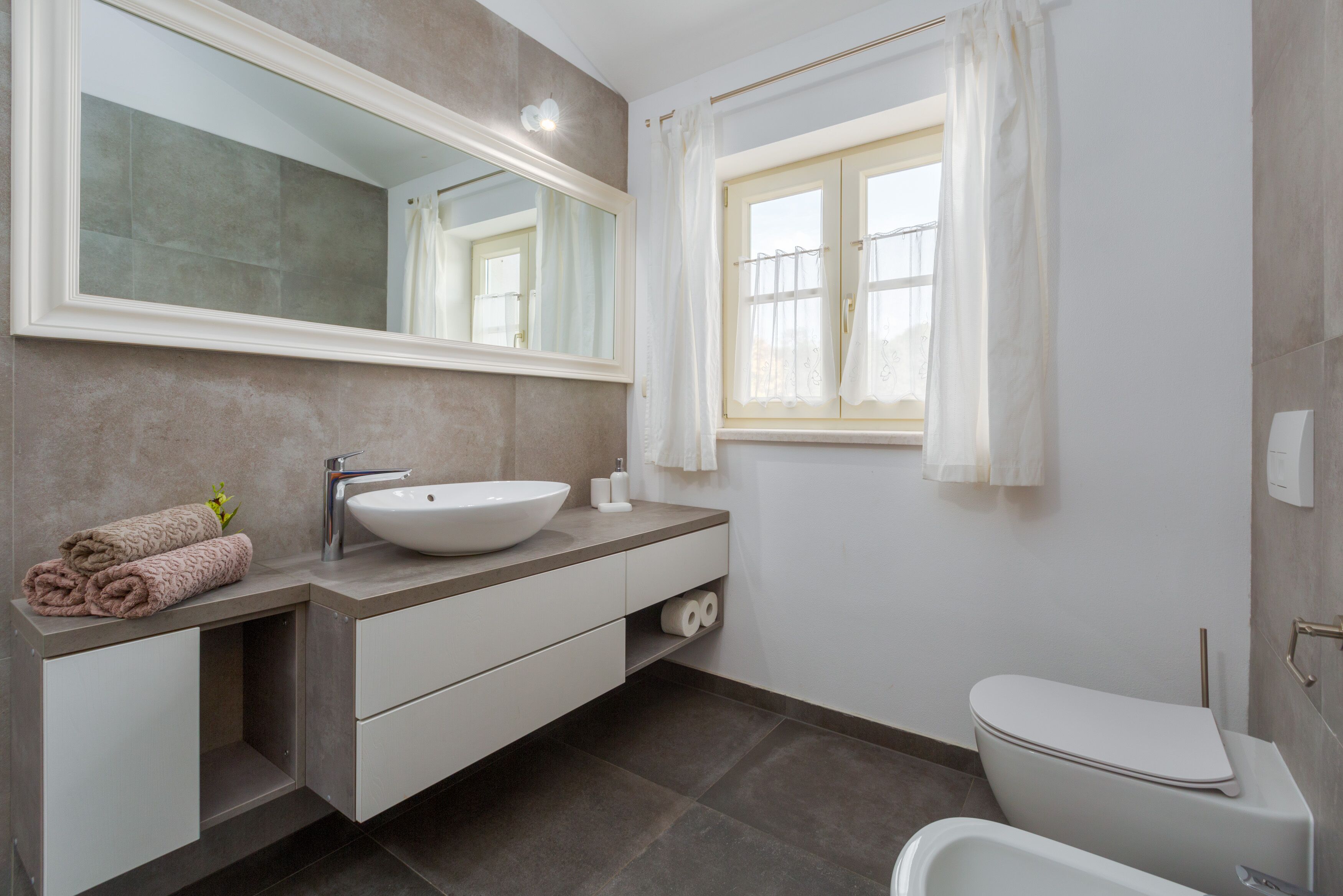 A modern bathroom with a large mirror, a sink, a toilet, and a window with curtains.