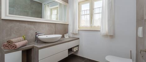 A modern bathroom with a large mirror, a sink, a toilet, and a window with curtains.