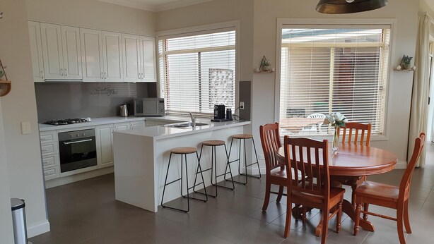 Una cocina moderna con armarios blancos, una isla central con taburetes de bar, una mesa de comedor con sillas de madera y un amplio ventanal con persianas.