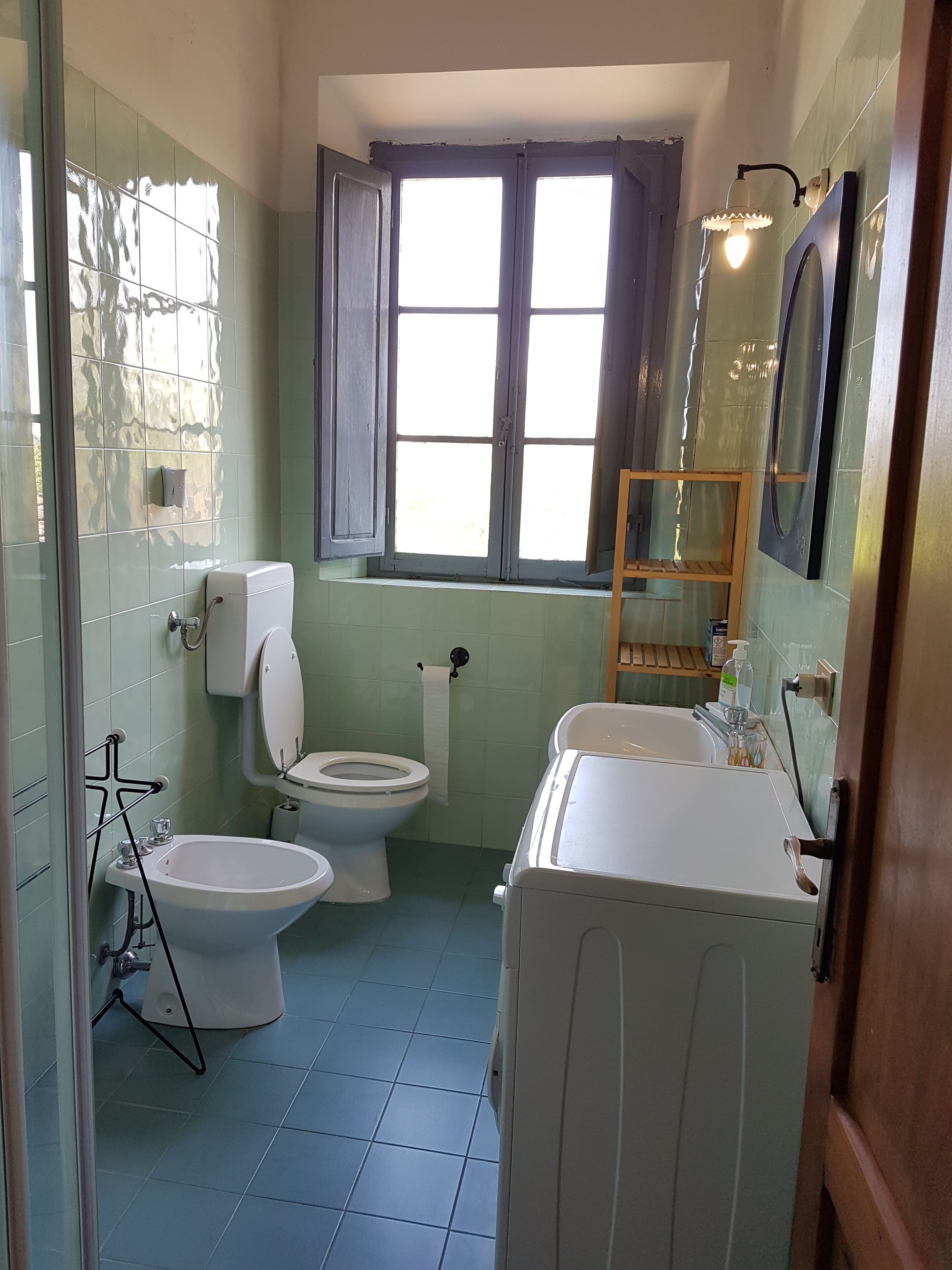 Ein Badezimmer mit Toilette, Bidet und Badewanne. Es gibt ein Fenster mit Vorhängen, einen Handtuchhalter und einen Spiegel.