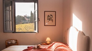 Ein Schlafzimmer mit einem Bett, einem Fenster, einem Nachttisch mit Lampe, einem gerahmten Bild an der Wand und Blick auf ein Feld.