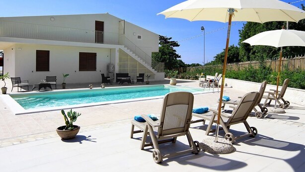 Un área junto a la piscina con sillones de descanso y sombrillas, una piscina y un edificio de dos pisos con balcón.