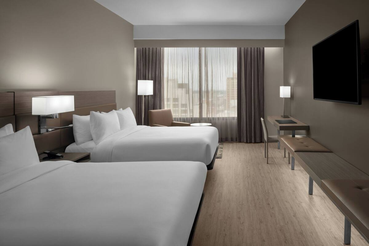 Une chambre d’hôtel avec deux lits, un bureau, une chaise, une télévision et une grande fenêtre avec des rideaux.