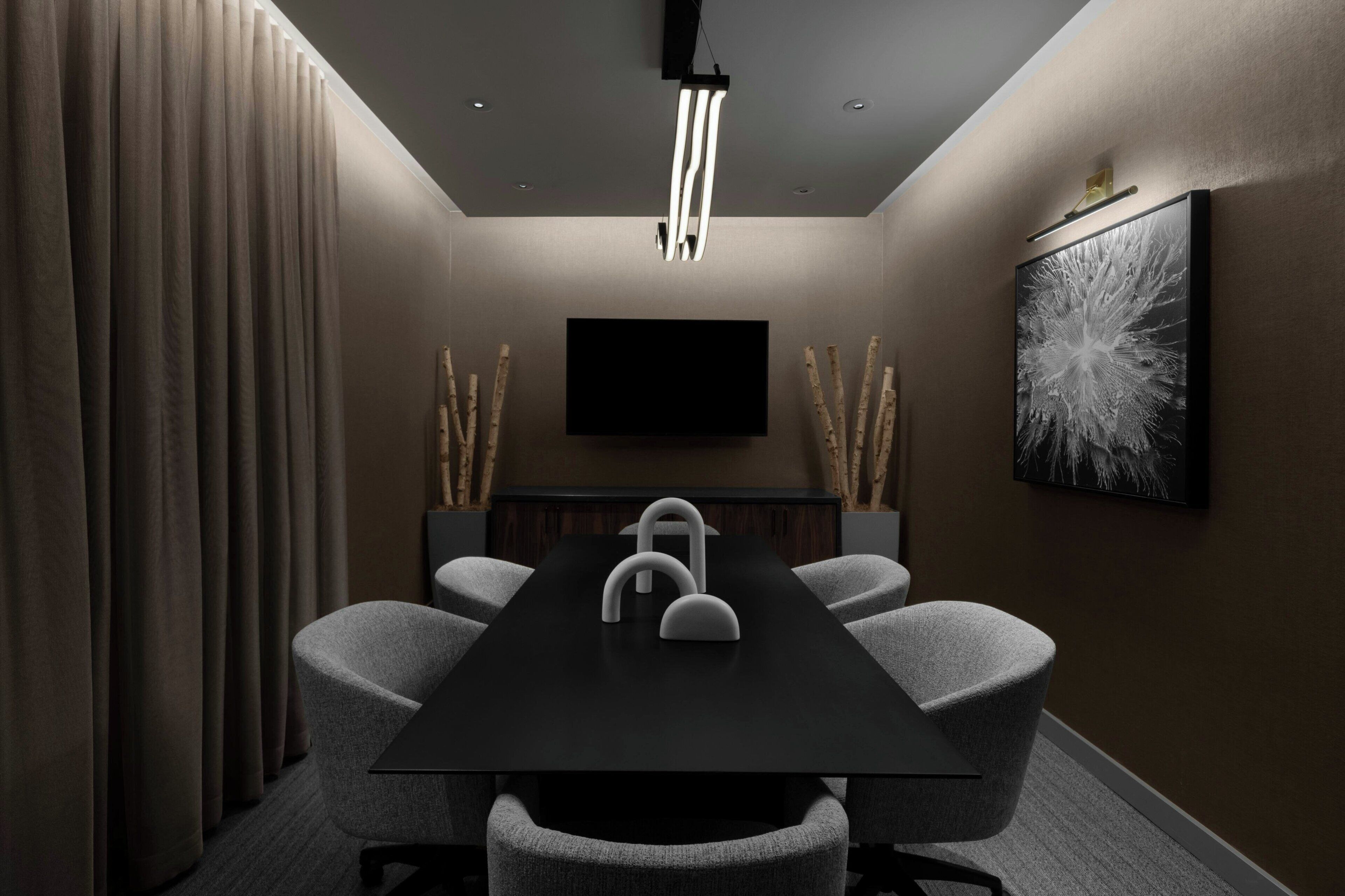 Uma sala de conferências com uma mesa preta, cadeiras cinzentas, uma TV instalada e uma luminária moderna.