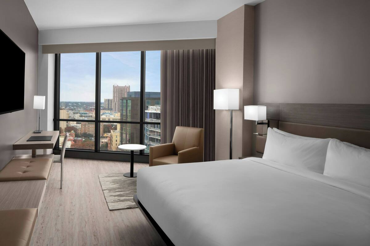 Une chambre d’hôtel avec un grand lit, un bureau, une chaise et une vue sur le paysage urbain grâce à de grandes fenêtres.