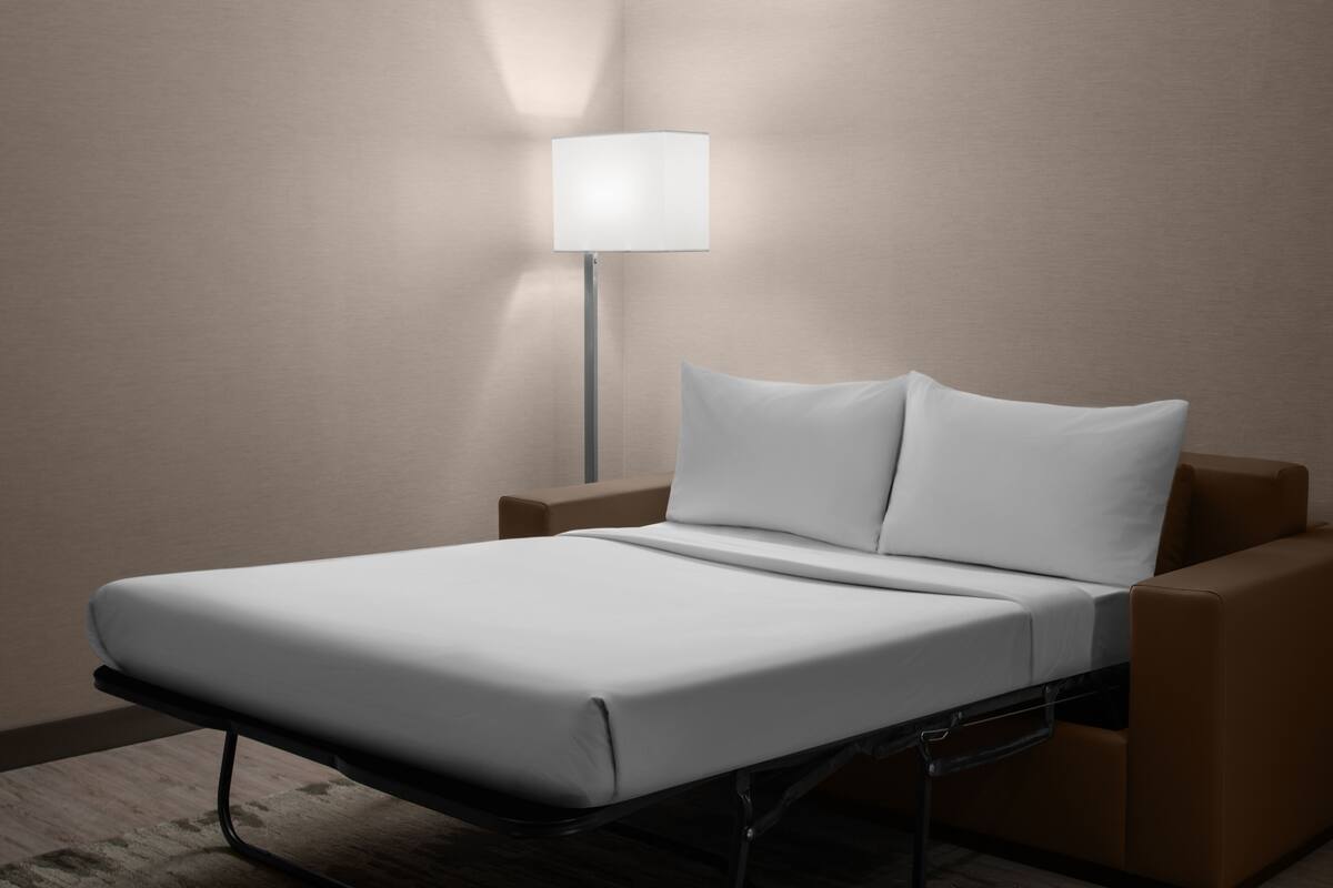 Un lit simple avec un matelas et des oreillers blancs, une table de chevet et une lampe sur pied, dans une pièce aux murs de couleur neutre et au plancher en bois.