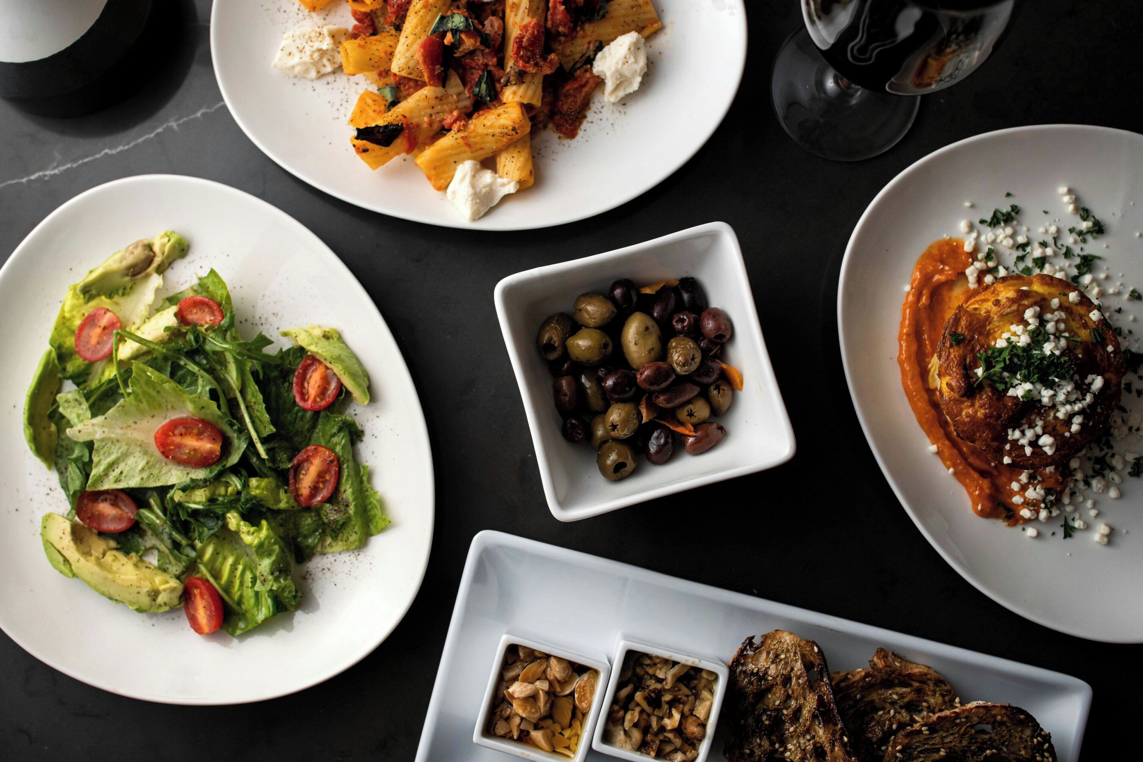 Una mesa con varios platos, como pasta, ensalada, aceitunas y carne a la parrilla.