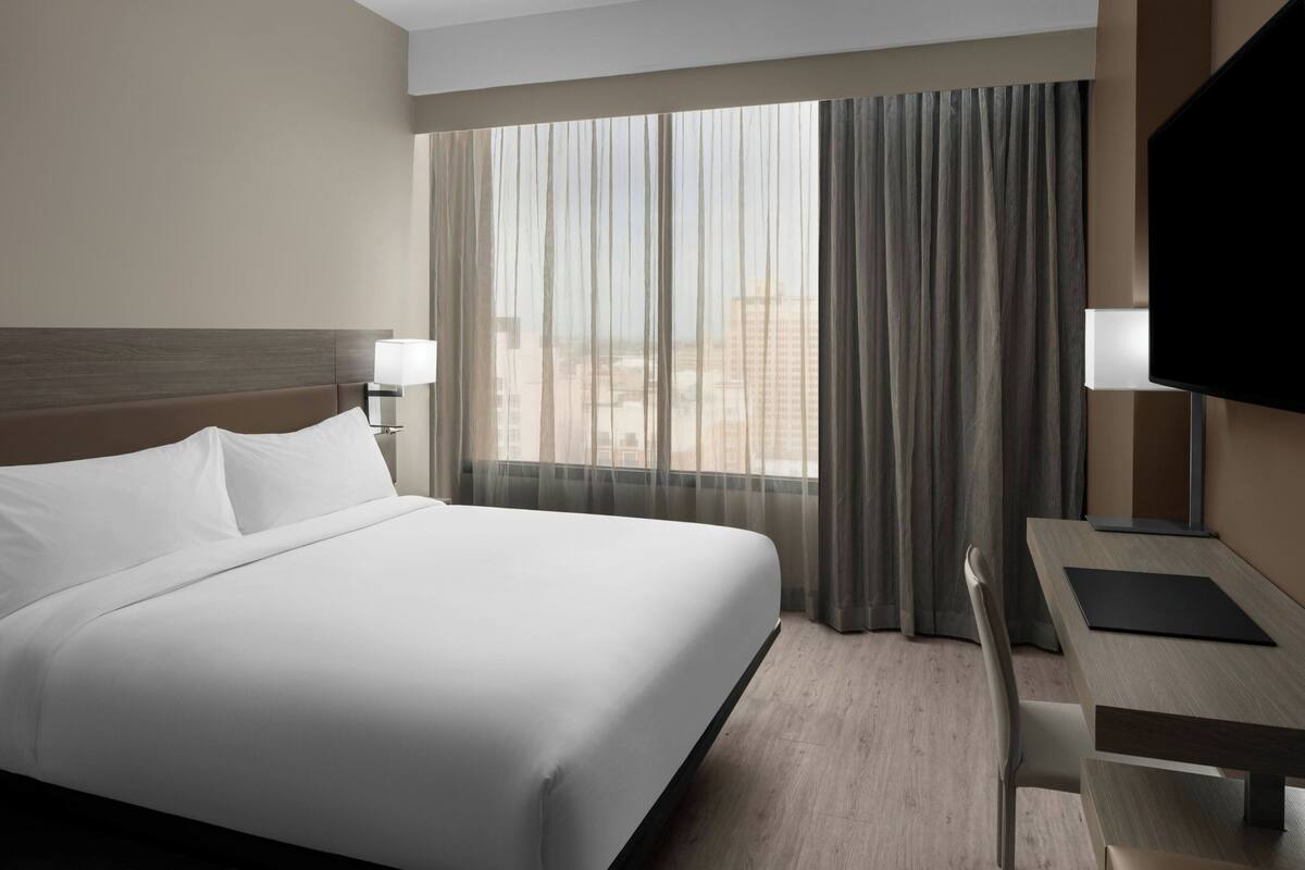 Une chambre d’hôtel avec un grand lit, un bureau, une chaise, une télévision et une fenêtre avec des rideaux.