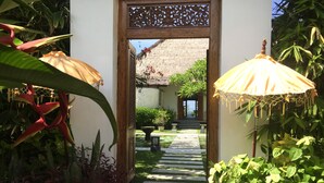 Un sentier menant à un bâtiment dont le toit est recouvert de chaume, entouré d’une végétation luxuriante et agrémenté d’un portail en bois décoratif.