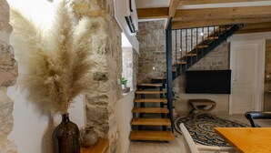 A narrow stone-walled corridor with a wooden staircase leading to a room. A large vase with dried plants is on the left, and a flat-screen TV is mounted on the wall in the room. - Villa Bante - Luxury Stone House (Kastela)
