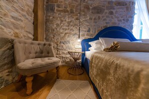 A bedroom with a stone wall, a blue headboard, a tufted armchair, a round side table, and a bed with white bedding. - Villa Bante - Luxury Stone House (Kastela)