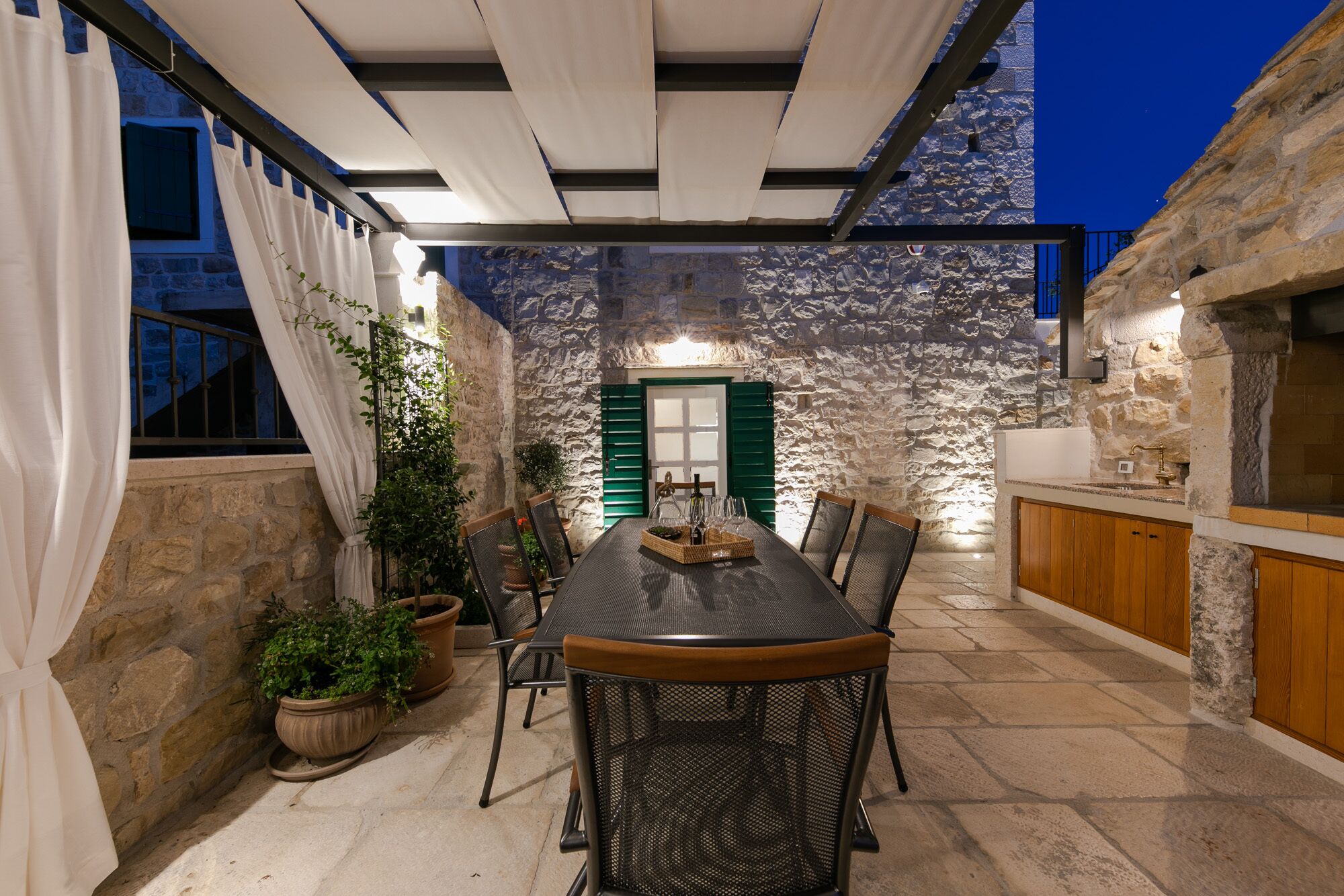 A covered outdoor dining area with a stone wall backdrop, a table with chairs, and a kitchen area with wooden cabinets.