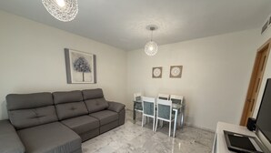 Un salon comprenant un canapé d’angle gris, une table à manger avec des chaises, un téléviseur à écran plat et un luminaire suspendu au plafond.