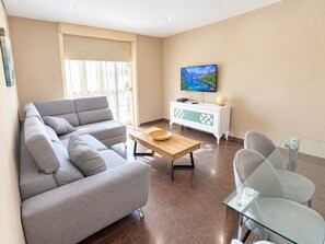 Ein Wohnzimmer mit grauem Sofa, einem Holztisch, einem Glastisch, einer weißen Kommode und einem Flachbildfernseher an der Wand.