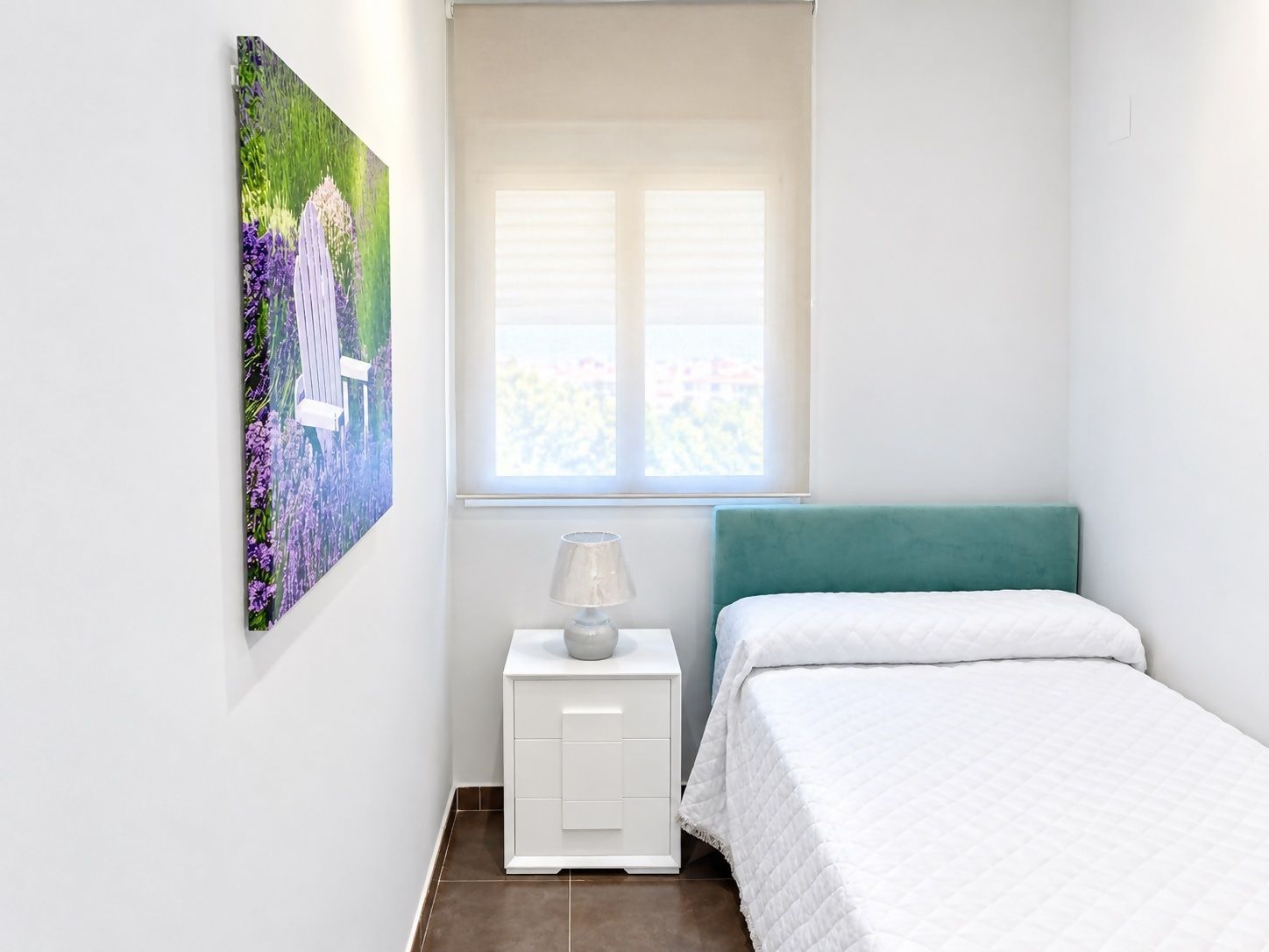 Una camera da letto con un letto, un comodino, una lampada, una finestra e un quadro appeso al muro.