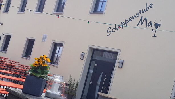 A building with a sign reading "Schoppenstube Ma" and a wine glass symbol. There are outdoor tables and chairs, and a potted plant with yellow flowers. - Schoppenstube May (Weigenheim)