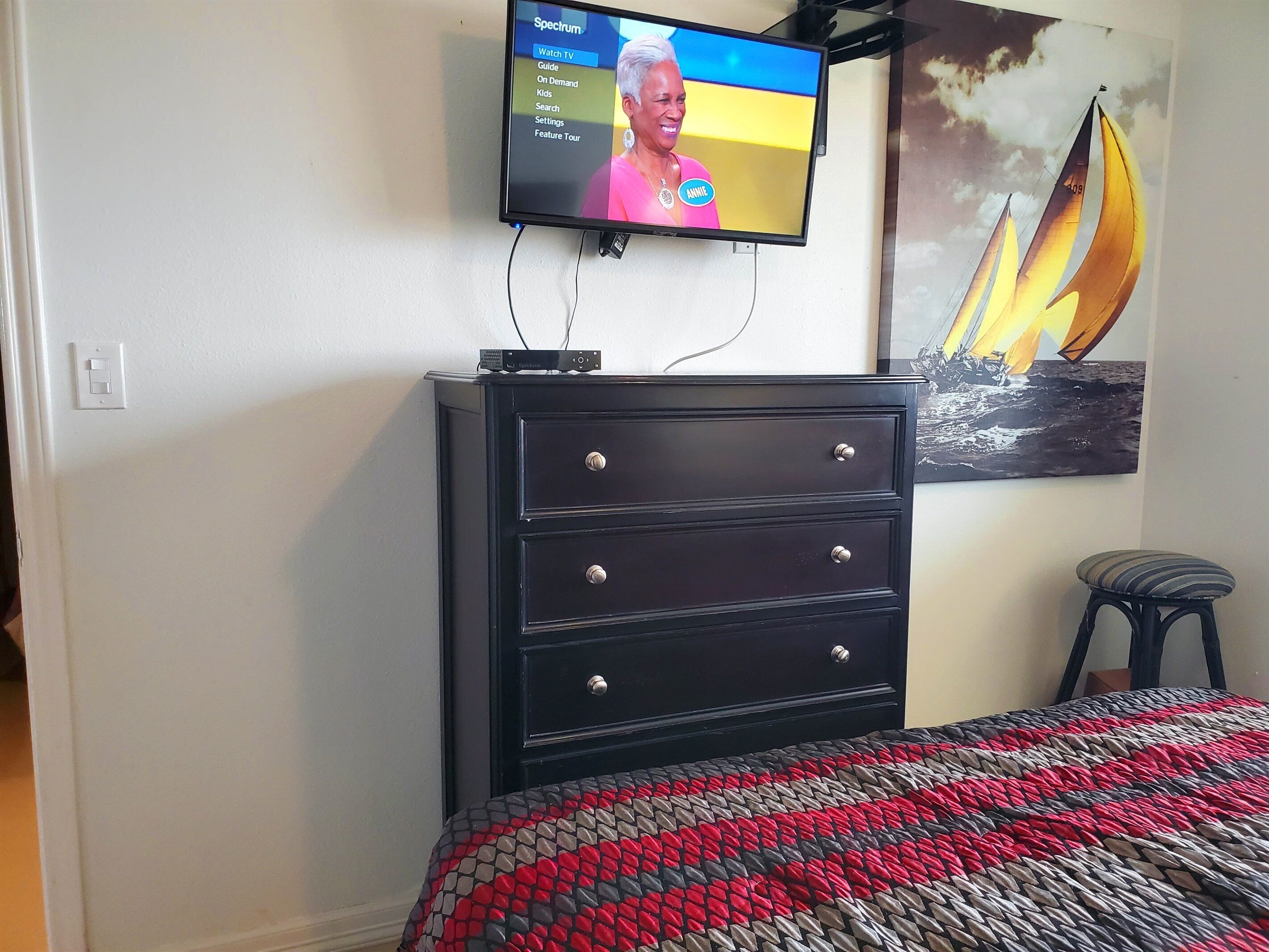 A bedroom with a bed, a dresser, a television mounted on the wall, and a painting of a sailboat.